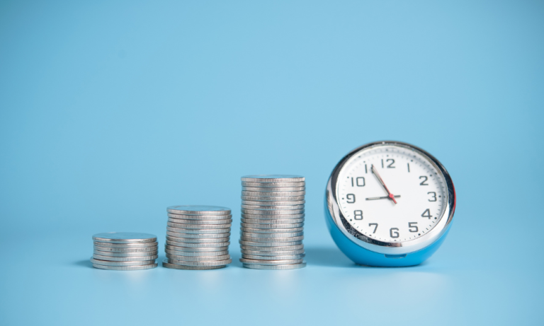 Against a pale blue background sits a stack of 7 10 pence coins, beside this stack sits a stack of 13 10ps, beside this sits a stack of 20 10ps. A small round silver clock sits next to the stacks of coins, it has a white face and displays the time 8:55